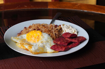 Gallo pinto, a typical Costa Rican dish with rice, red beans, sausage, cream, and a fried egg, and a glass of coffee on a table with a nature background.