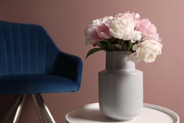 Beautiful peonies in vase on white table and armchair near pink wall