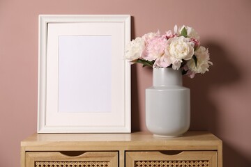 Beautiful peonies in vase and picture frame on wooden dresser near pink wall. Space for text
