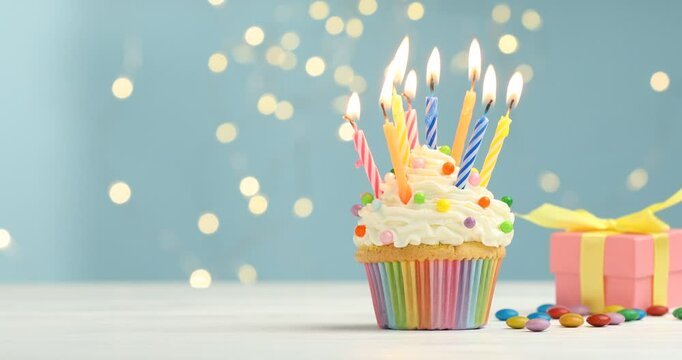 Burning candles in cupcake, gift and candies on table against blue background with blurred lights