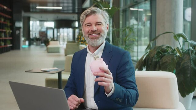 Happy mature businessman entrepreneur save money using pink piggybank putting dollar cast inside. Success, economy, investment, deposit. Bearded guy in suit sitting with laptop in modern office lobby.