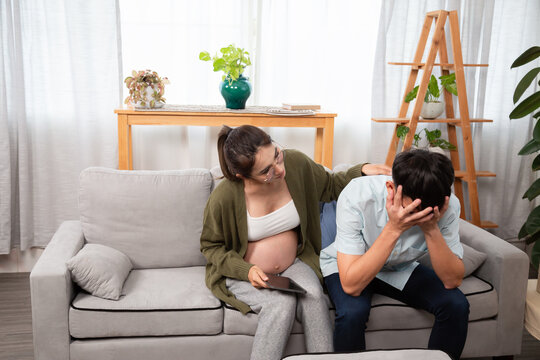 Pregnant woman sitting on couch, comforting stressed boyfriend holding head in hands, depicts emotional support, job loss stress, and challenges faced by expecting parents.