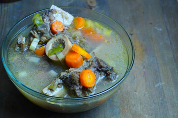 A close-up of a hearty bone marrow soup, featuring tender meat, vibrant carrots, and green vegetables in a clear glass bowl on a rustic wooden table.