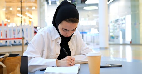 Focused muslim student girl in hijab is making notes in notebook, sitting in cafe at shopping mall food court, studying and drinking coffee. Muslim sign a contract, conducts business Arab woman