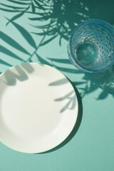 Empty white plate with glass of water on the blue background with palm leaves shadows. Summer table settings. Copy space