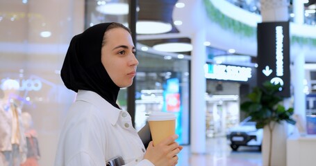 Young muslim businesswoman in hijab enjoying coffee break, working on tablet while walking through modern shopping mall interior