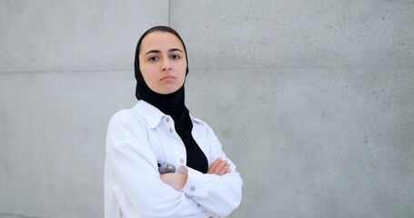 Serious young muslim professional wearing hijab and white shirt, confidently standing against gray wall with arms crossed and direct gaze