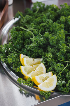 Close-up of fresh curly parsley with lemon wedges on a metal tray, ready for food preparation or garnish.

