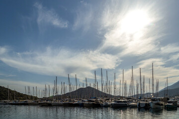 Obraz premium Many sailboats with tall masts docked at marina pier, backlit by sun. Reflections on water, blue sky, green hills in background. Kas, Mediterranean, Turkey.