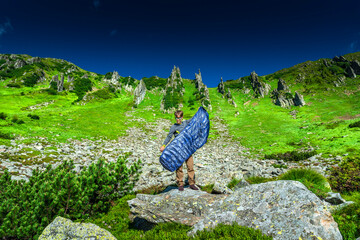 Backpacker airing out a sleeping bag on rocky mountain terrain under a clear blue sky. Summer...