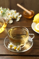 Delicious chamomile tea in glass cup, fresh flowers, lemon and honey on wooden table, closeup
