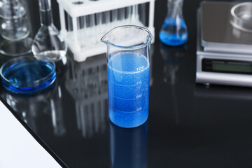 Beaker with blue liquid, other laboratory glassware and scale on black mirror table, closeup