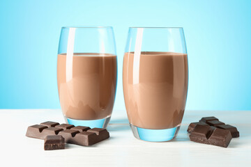 Tasty chocolate milk in glasses and pieces on white wooden table against light blue background
