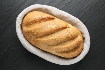Loaf of bread with sesame seeds in basket on black textured table, top view