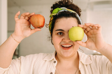 Portrait of a young smiling curly-haired plus-size woman with apple in her hands.A woman closes her eye with one apple.Healthy eating and healthy lifestyle.