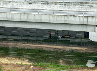 A lone construction worker in a yellow helmet walks beneath a highway bridge during the day,...