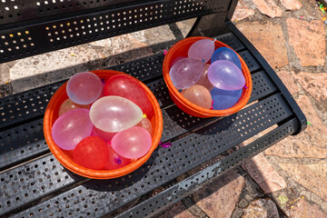 Two bright orange bowls filled with colorful water balloons sit on a bench, prepared for a fun and messy outdoor water balloon fight in the park.