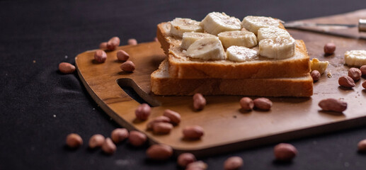 Delicious toast topped with peanut butter, banana slices,Crunchy bread slice with creamy peanut butter and sliced banana, arranged artfully on a clean chopping board with peanuts scattered arround it
