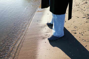 A person stands at the edge of the water on a sandy beach, wearing light-colored pants and shoes