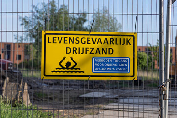 A stark yellow Dutch sign warns of drijfzand "Lethal Quicksand" and forbidden access, mounted on a fence, emphasizing danger and safety.