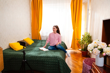 Young woman sitting on her bed and recording a video, creating engaging content for her social media followers. Wide shot