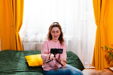 Portrait of enthusiastic content creator sitting on a bed and filming an engaging video for her...