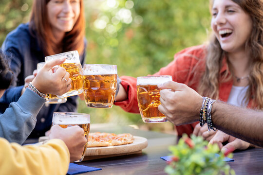 Friends toasting with beer mugs and eating pizza at an outdoor pub garden party in summer.
