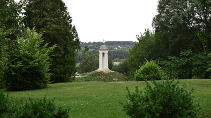 Obraz premium a small bell tower in the park. white bell tower surrounded by green trees. religious premises, Christianity or Catholicism. stone walls, cross on the dome. spring or summer time