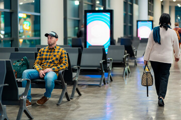 Obraz premium Wide shot of adult Caucasian man in cap and jacket using smartphone in airport waiting area sitting in chair. Concept of boarding at terminal before flight