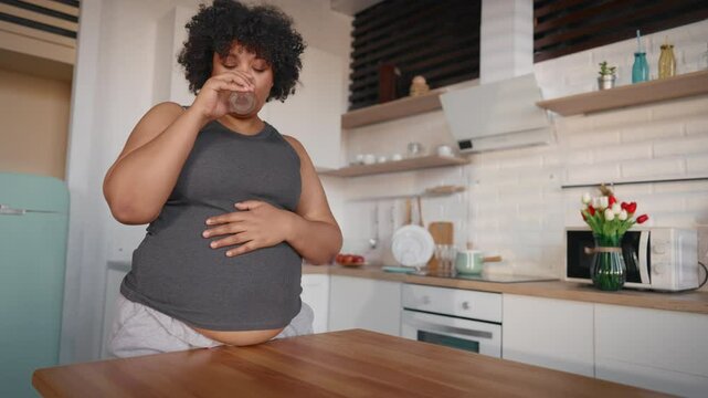 Pregnant woman feeling tiredness in morning, drinking water in home kitchen. Portrait of pretty young adult black lady with big belly, eight month of pregnancy, bad feeling and nausea, motherhood