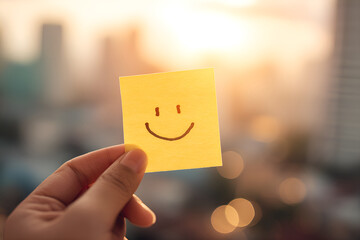Hand holding sticky note with smiley face at sunset in city