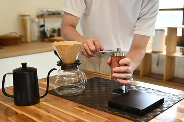 A man is grinding coffee beans in a coffee grinder. The coffee grinder is on a wooden table.