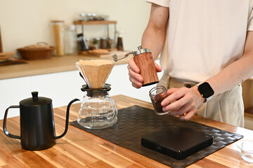 A man is grinding coffee beans in a coffee grinder. The coffee grinder is on a wooden table.