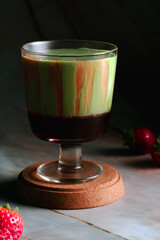 A glass of a strawberry matcha latte, showcasing distinct layers of dark red, light cream and vibrant green with a strawberry on the side.