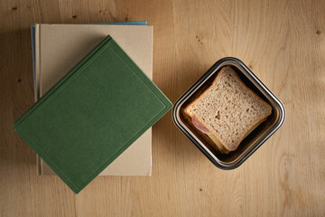 A table top view of green hardback book lies beside a metal lunch box containing a sandwich, set on a wooden surface for a school or study scene. Back to school