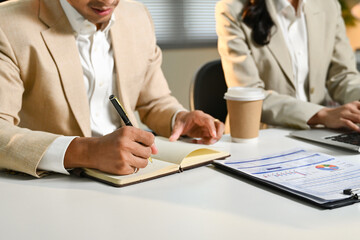 Two people in suits are sitting at a desk with a notebook and a laptop. One of them is writing in the notebook while the other is typing on the laptop.