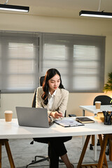A woman in a business suit is sitting at a desk with a laptop and a stack of papers. She is focused on her work