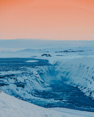 The majestic Gullfoss waterfall in Iceland, partially frozen in winter, showcases the dramatic power of nature amidst a snowy landscape and a vibrant twilight sky.