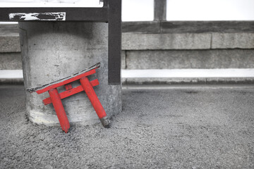 A small, broken red torii gate leans against a concrete pillar on a gray asphalt surface. 