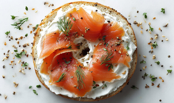 A bagel with cream cheese and salmon garnished with dill on a white surface with seeds around it