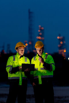 Vertical image of petrochemical workers or technicians using laptop to work at night and stand in front of the factory with light.