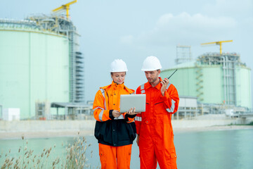 Two petrochemical or gas factory workers man and woman stand in front of factory tank near the bay and work together using laptop.