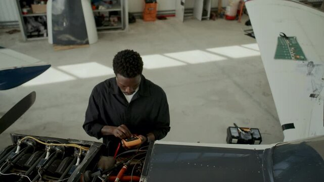 African American mechanic operating digital multimeter while testing aircraft engine components inside maintenance hangar. High angle view
