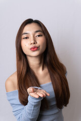 Fototapeta premium Portrait of graceful, sensual blonde Asian woman wearing fashionable light blue off-shoulder top with passionate face blowing towards camera isolated on white background. Beauty, fashion concept.