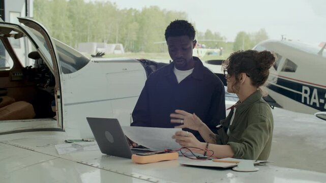 African American male mechanic and his Caucasian female colleague discussing technical documents and inspecting repair plans beside small aircraft in hangar