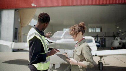 African American ground technician and Caucasian female inspector discussing condition of small aircraft in front of open hangar at airfield - Powered by Adobe