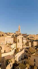 Naklejka premium Aerial view of Matera, Italy, bathed in golden morning light. The iconic stone dwellings and cathedral tower rise above the canyon, showcasing this UNESCO-listed historic wonder