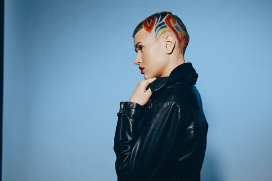 Fototapeta woman with colorful geometric shaved hairstyle wearing black leather jacket stands against soft blue background in dramatic side profile exhibiting bold modern style