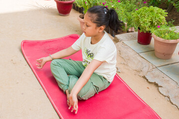 A Indian young girl sitting in Sukhasana (Easy Pose) on a red yoga mat, focusing on mindfulness and breathing. The child is practicing yoga outdoors. kids' yoga, physical fitness, and mental wellness.