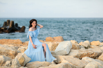 Beautiful young girl with long dark hair in a blue long dress on the beach at sea in summer